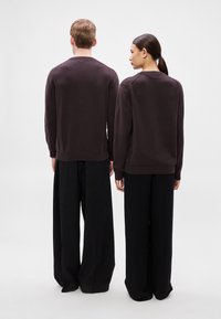 Duas pessoas a usar camisolas escuras e largas e calças pretas largas, em pé costas voltadas uma para a outra contra um fundo simples.