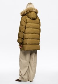 Abrigo acolchado color caqui con capucha con ribete de piel, diseño acolchado horizontal y ajuste holgado, combinado con pantalones de pierna ancha color beige claro.