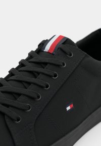Scarpa da ginnastica nera in tela, con lacci piatti, suola in gomma e una linguetta multicolore con strisce rosse, bianche e blu.