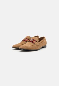 Mocassini in suede color sabbia con design a punta, caratterizzati da una fascia decorativa con accenti rossi e neri e un emblema dorato sul davanti.