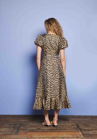 Mujer con cabello rizado que lleva un vestido midi de estampado de leopardo con mangas abullonadas y dobladillo con volantes, de pie sobre un suelo de madera contra una pared azul pálido.
