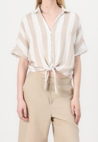 Camicia a maniche corte in lino con righe beige e bianche, chiusura con bottoni e orlo annodato. Abbinata a pantaloni ampi beige chiaro.