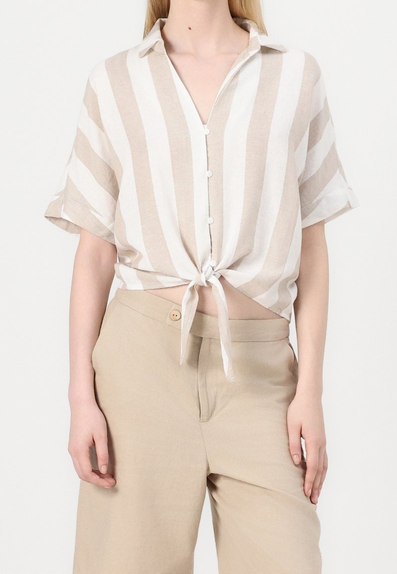 Camicia a maniche corte in lino con righe beige e bianche, chiusura con bottoni e orlo annodato. Abbinata a pantaloni ampi beige chiaro.