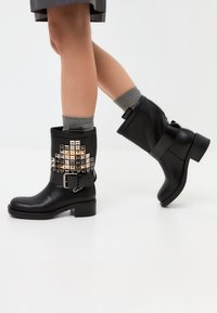 Botines negros hasta el tobillo con un acabado texturizado, adornados con acentos metálicos de tachuelas, una correa ancha y un tacón bajo en bloque.