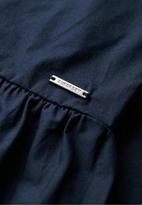 Marineblauer Stoff mit geraffter Textur, versehen mit einem silbernen Metallschild, das "SUPERDRY" eingraviert hat, um das Branding zu kennzeichnen.