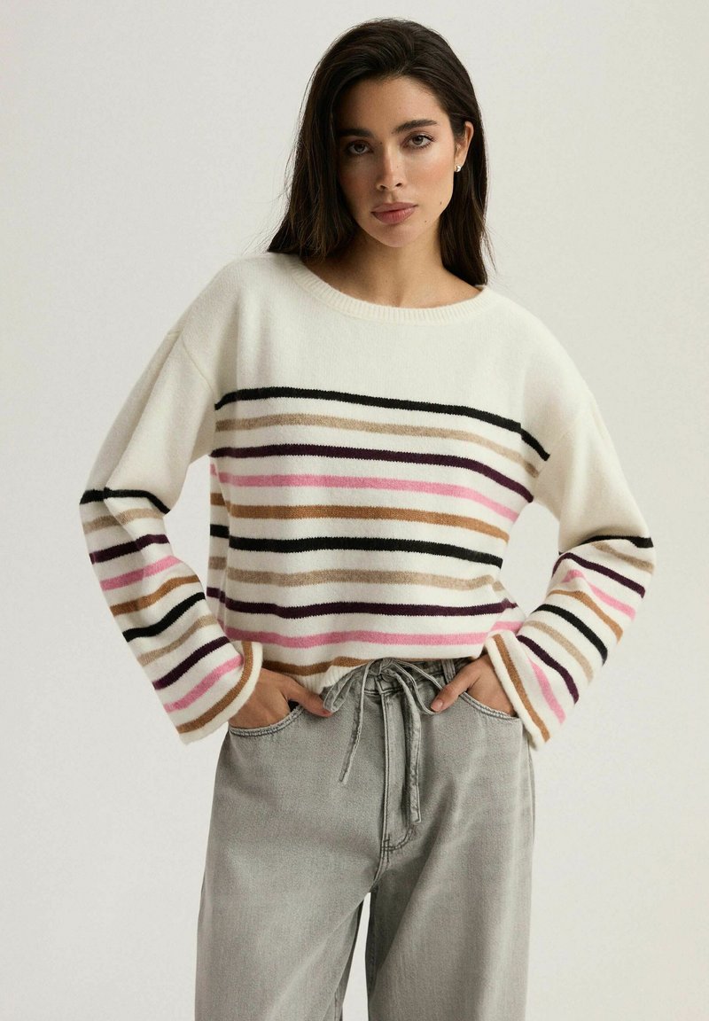 Sweter w paski w kolorze kremowym z poziomymi paskami w kolorze czarnym, różowym i beżowym. Posiada okrągły dekolt i luźne rękawy. Szare spodnie.