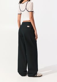 Vrouw staat met haar rug naar ons gekeerd, draagt zwarte wijde broek, een doorschijnend wit T-shirt met zwarte randen en witte sneakers.