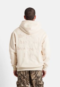 Sudadera con capucha crema que presenta una cita bordada en la parte posterior, con puños acanalados y un corte relajado. Combinada con pantalones cargo de camuflaje.