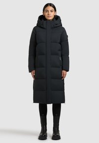 Žena stojící vzpřímeně, oblečená v dlouhém černém péřovém kabátu s kapucí, černých legínách a černých kotníkových botách na světle šedém pozadí.