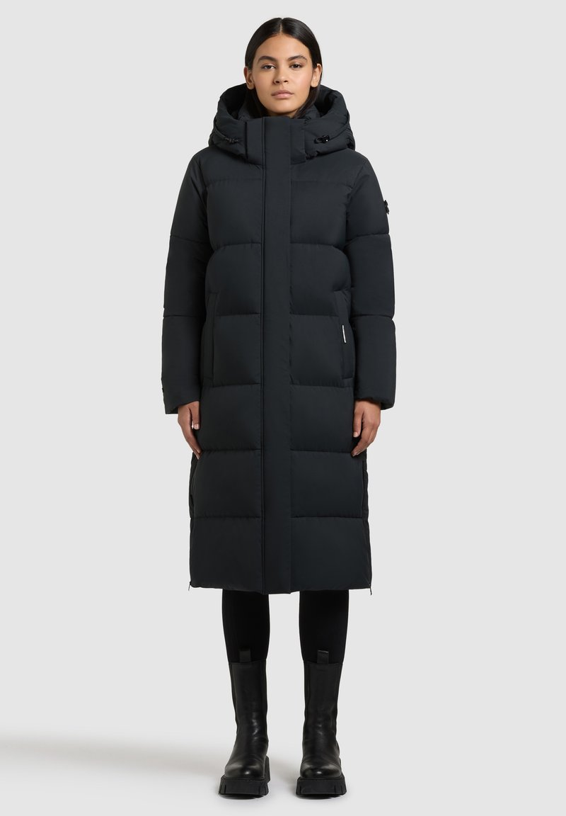 Žena stojící vzpřímeně, oblečená v dlouhém černém péřovém kabátu s kapucí, černých legínách a černých kotníkových botách na světle šedém pozadí.