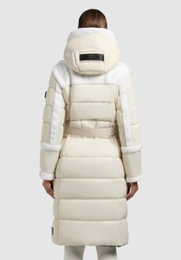 Beige puffrová bunda s bílými umělými kožešinovými akcenty a páskem v pase. Má velkou kapuci a logo nášivku na zadní straně. Texturované panely po celé ploše.