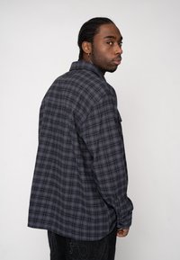 Grijze geruite blouse met lange mouwen en knoopsluiting, met een relaxte pasvorm en rechte zoom, gemaakt van zacht stof. Achteraanzicht toont het gestructureerde materiaal.