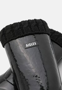 Bottes en caoutchouc noires imperméables avec un bord en tricot noir texturé. Présente une étiquette de logo rectangulaire avec la marque "AIGLE 1853."