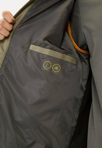 Waterafstotende jas met een interieur in donkergrijs, voorzien van een zak met een beige band. Labels geven iconen voor water- en vlekbestendigheid aan.