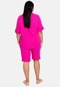 Chemise courte à manches courtes en lin rose vif et shorts assortis arrivant aux genoux, avec des manches retroussées et une coupe ample, portés avec des sandales beiges.