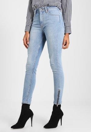Jeans skinny de mezclilla azul claro con cintura alta, que cuentan con cremalleras en los tobillos y un ligero desgastado. Combinados con botas negras hasta el tobillo y una blusa a rayas.