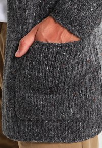 Cárdigan de punto gris con una superficie texturizada, que presenta un gran bolsillo en la parte frontal, combinado con pantalones beige, destacando su diseño casual.