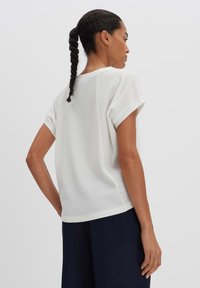 Top bianco a maniche corte con texture, orli arrotolati, vestibilità rilassata e fondo arrotondato, abbinato a pantaloni scuri. Vista posteriore.
