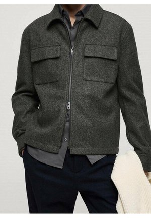 Homme portant une veste zippée gris foncé avec des poches à rabat sur la poitrine par-dessus une chemise grise, tenant un pull léger en maille dans une main.