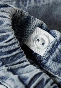 Džinsinio audinio artimas vaizdas, kuriame matyti tekstūruotas mėlynas raštas. Centre yra matoma balta etiketė su aiškia sagos detale.