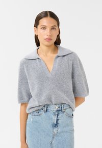 Sivi pulover s kratkimi rokavi in V-izrezom ter ovratnikom, izdelan iz teksturiranega mehkega materiala, v kombinaciji z svetlo modrim džins krilom.