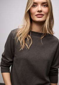 Pull gris foncé à large encolure, poignets côtelés et texture subtile, porté par une personne aux longs cheveux châtain clair.