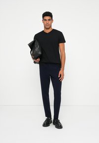 Black V-neck T-shirt, navy slim-fit trousers, and black leather shoes. Model holds a black leather bag, standing against a plain background.