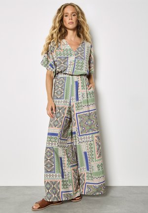 Vrouw met lang, golvend haar die een jumpsuit met korte mouwen en patroon draagt in groene, blauwe en beige tinten, staande met één hand in haar zak.