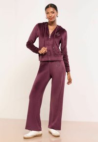 Burgundská velúrová mikina na zips s vreckami a malým zdobeným logom, spárovaná s ná matching širokými nohavicami a bielymi teniskami. Ploský povrch.