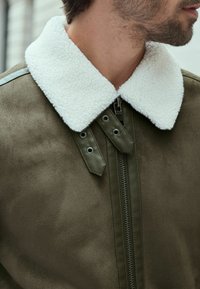 Chaqueta verde oliva con un cuello blanco y esponjoso y cierre de cremallera. Presenta herrajes de metal y una superficie lisa y texturizada.