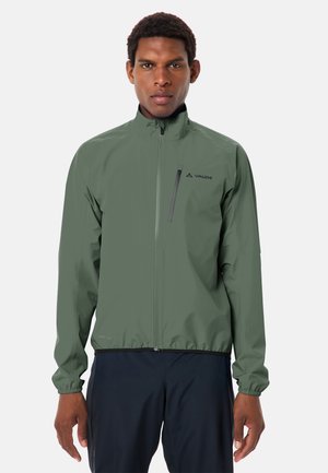 Uomo che indossa una giacca verde oliva con cerniera e tasca sul petto, pantaloni neri, in piedi davanti a uno sfondo chiaro uniforme.