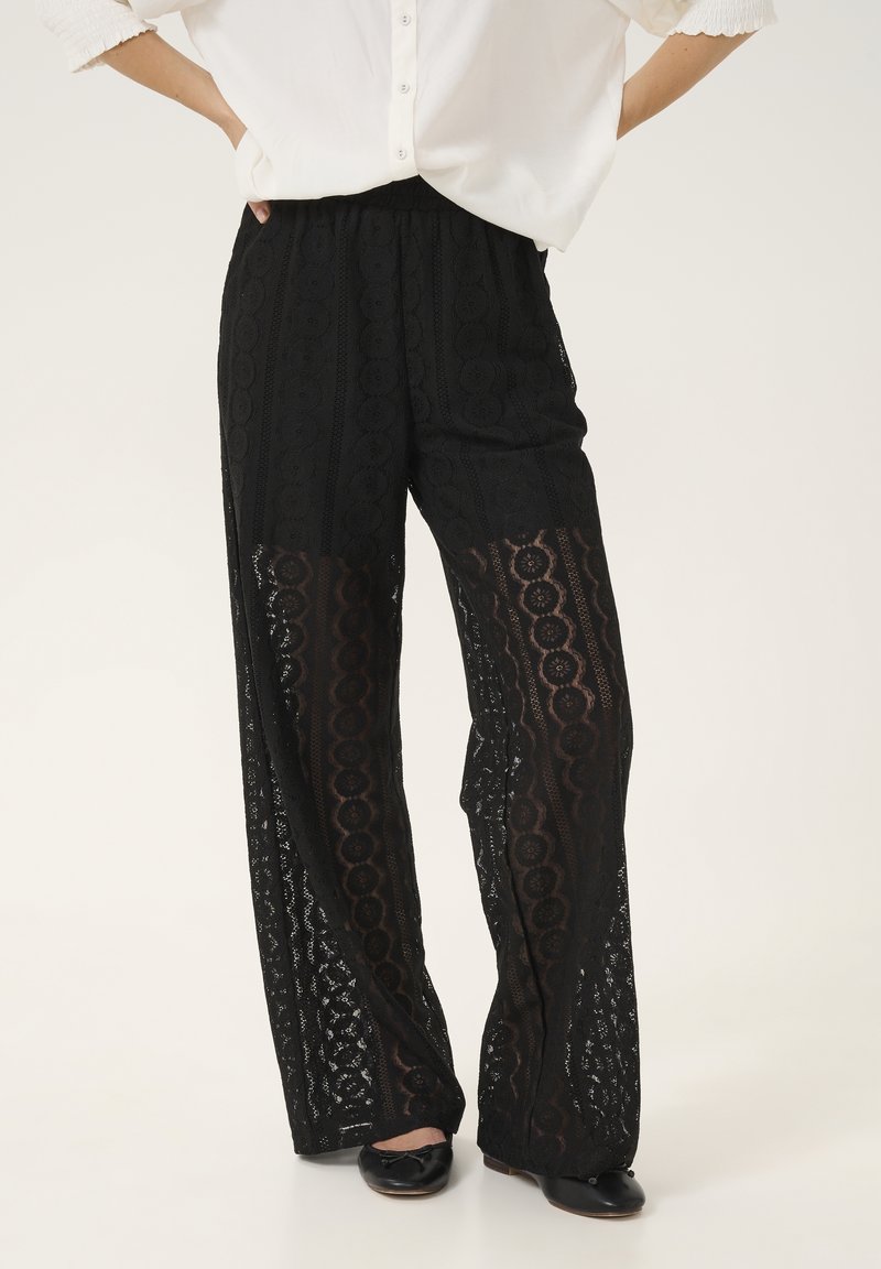 Femme portant un pantalon large en dentelle noire à motifs floraux et des ballerines noires, associée à une chemise blanche boutonnée.