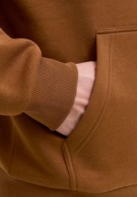 Hånden er delvist inde i forlommerne på en brun sweatshirt med ribbet manchet og synlige sømme.