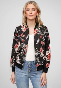 Veste bomber en tissu noir ornée de fleurs rouges et blanches. Elle présente des poignets côtelés et une fermeture éclair, associée à un jean taille haute bleu clair.