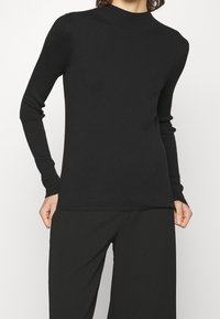 Haut noir à col montant en côtelé, à manches longues ajustées, avec une texture lisse et des détails de couture subtils, porté avec un pantalon noir à jambes larges.