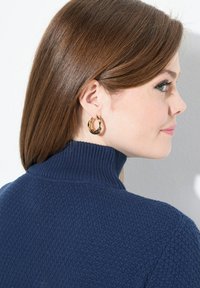 Frau mit geraden braunen Haaren, die goldene Creolen trägt und einen navyblauen, strukturiertenRollkragenpullover anhat, seitlich blickend.