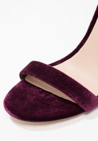 Sandale à talon en velours de couleur bourgogne foncé avec une large bride avant et une semelle beige. Sa texture douce et sa forme élégante renforcent son design.