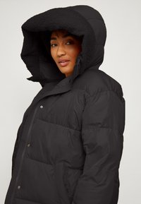 Schwarze Daunenjacke mit einer großen, gefütterten Kapuze, Druckknopfverschluss und Seitentaschen. Verfügt über ein gestepptes Design und eine weiche Textur.