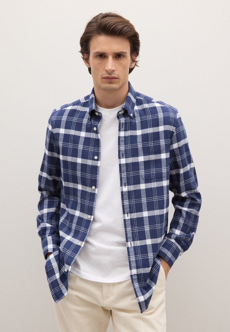 Blau-weiß kariertes Hemd aus einem weichen Material, mit einem Button-Down-Kragen, langen Ärmeln und einer Brusttasche. Getragen über einem weißen T-Shirt.