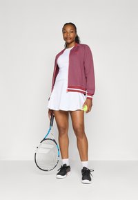 Bordo sportinė striukė su dryžuotais rankovėmis, balta teniso suknelė, juodi sportbačiai ir teniso raketė. Laiko geltoną teniso kamuoliuką.