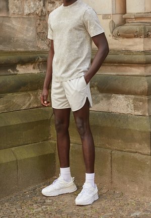 T-shirt texturé gris clair, shorts crème avec un motif en losanges et baskets blanches. Le mannequin se tient devant un mur en pierre, les mains dans les poches.