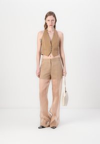 Väldigt kort brunt väst med knappar, matchat med genomskinliga beige vida byxor. Modellen håller en liten krämfärgad handväska och står mot en vit bakgrund.