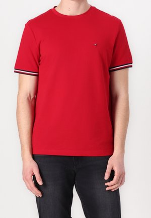 Mann trägt ein rotes T-Shirt mit kurzen Ärmeln, gestreiftem Saum an den Ärmeln und kleinem Logo auf der Brust, kombiniert mit schwarzen Hosen.