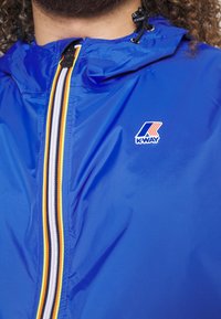 Gros plan d'une veste K-Way bleue avec fermeture éclair rayée multicolore et cordons de capuche, portée par une personne aux cheveux bouclés et à la barbe.
