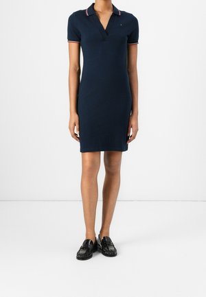 Schmal geschnittenes marineblaues Polokleid mit kurzen Ärmeln und rot-weißem Besatz am Kragen und an den Ärmelabschlüssen, kombiniert mit schwarzen Loafers.