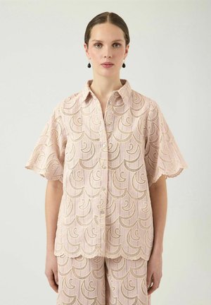 Femme aux cheveux plaqués en arrière, portant un chemisier en dentelle rose pâle à manches courtes et un pantalon assorti avec des bords festonnés et une broderie incurvée.