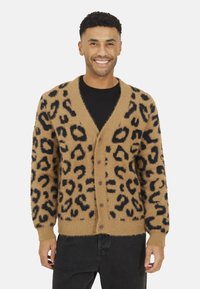 Cardigan à imprimé léopard en beige avec des taches noires. Texture douce et duveteuse, décolleté en V, fermeture à bouton, et poignets côtelés. Porté par-dessus un t-shirt noir.