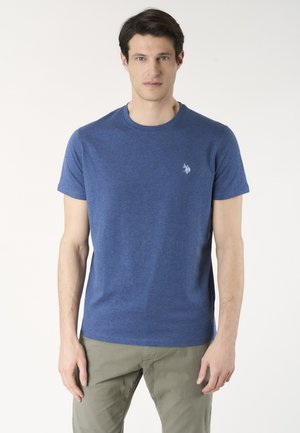 Uomo che indossa una maglietta blu a maniche corte con un piccolo logo sul petto e pantaloni verde oliva, in piedi davanti a uno sfondo bianco.