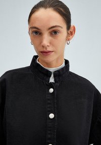 Zwarte denim jas met hoge kraag, voorzien van een zilveren knoopsluiting en een gestructureerde afwerking. Strak ontwerp, geen patronen of versieringen.
