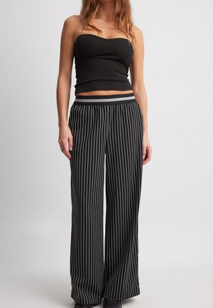 Vrouw met een zwarte strapless top en zwarte hoge taille broek met wijde pijpen, voorzien van witte verticale strepen en een gestreepte tailleband.